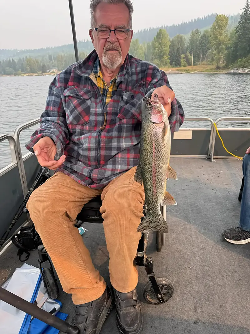 A guest in a wheelchair holding a large wild rainbow trout on a boat on Quesnel Lake